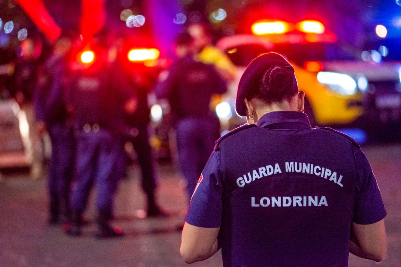 A municipal guard stands on duty in the vibrant streets of Londrina at night.