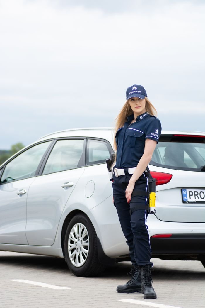 Female police officer standing confidently beside a white patrol car on a sunny day.
